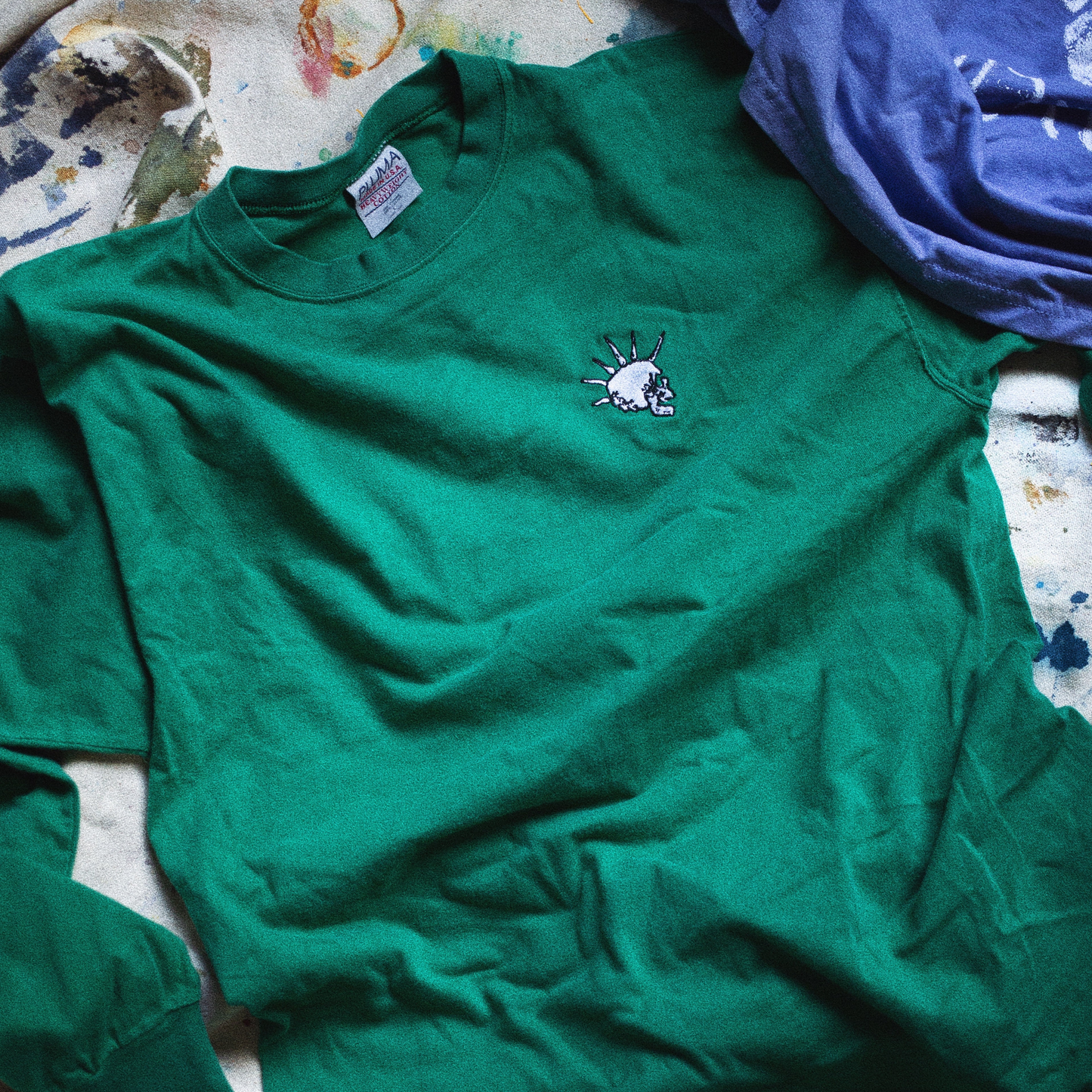 Green longsleeve shirt with skull logo on a canvas with colorful stains and paint