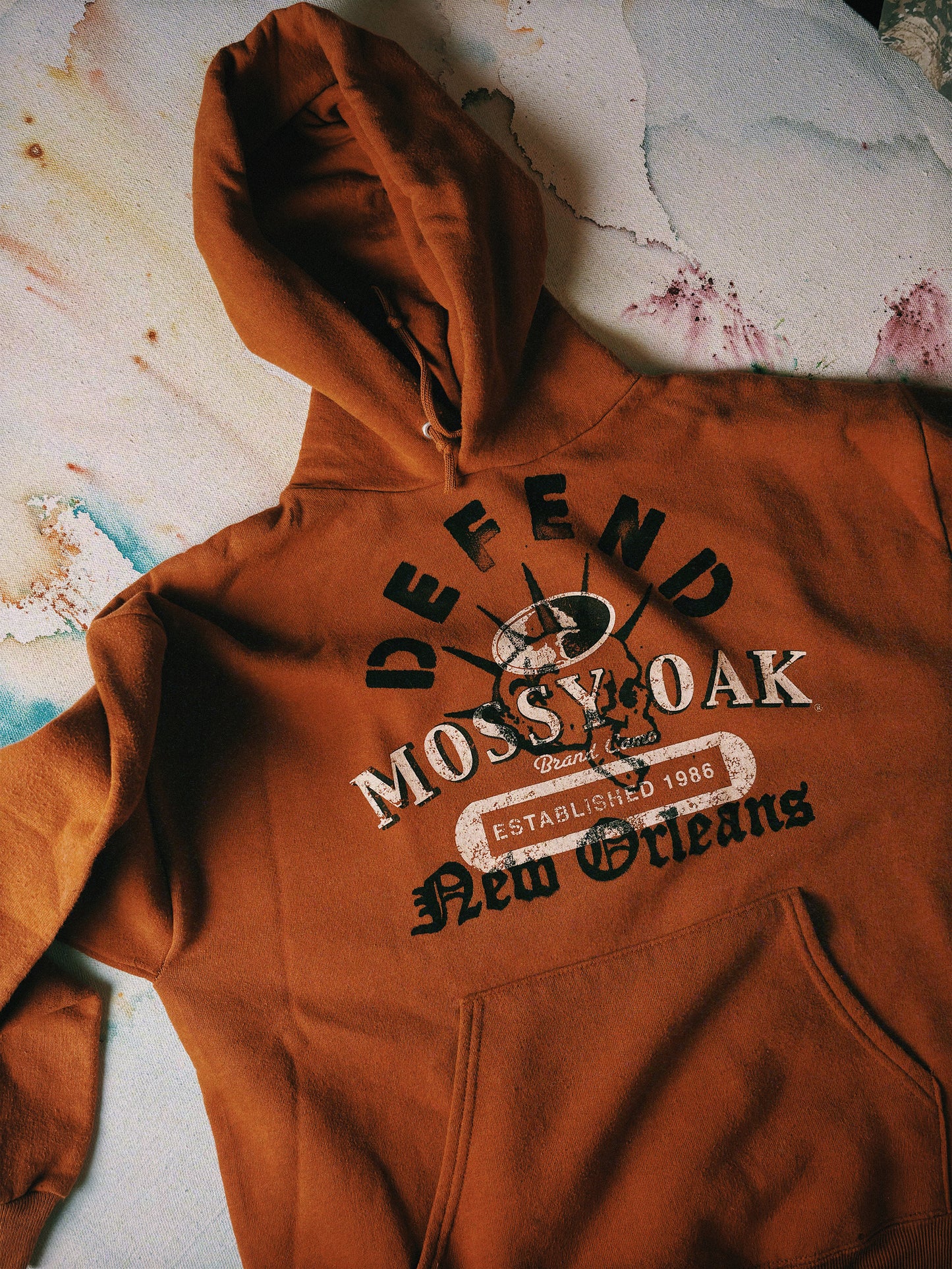 Brown hoodie with 'Defend Mossy Oak New Orleans' text on a textured surface