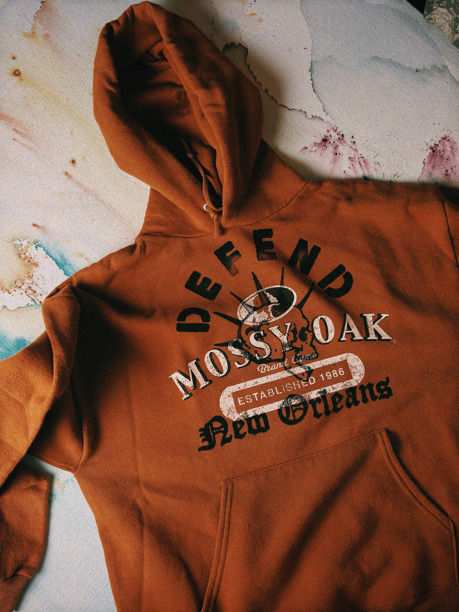 Brown hoodie with 'Defend Mossy Oak New Orleans' text on a textured surface